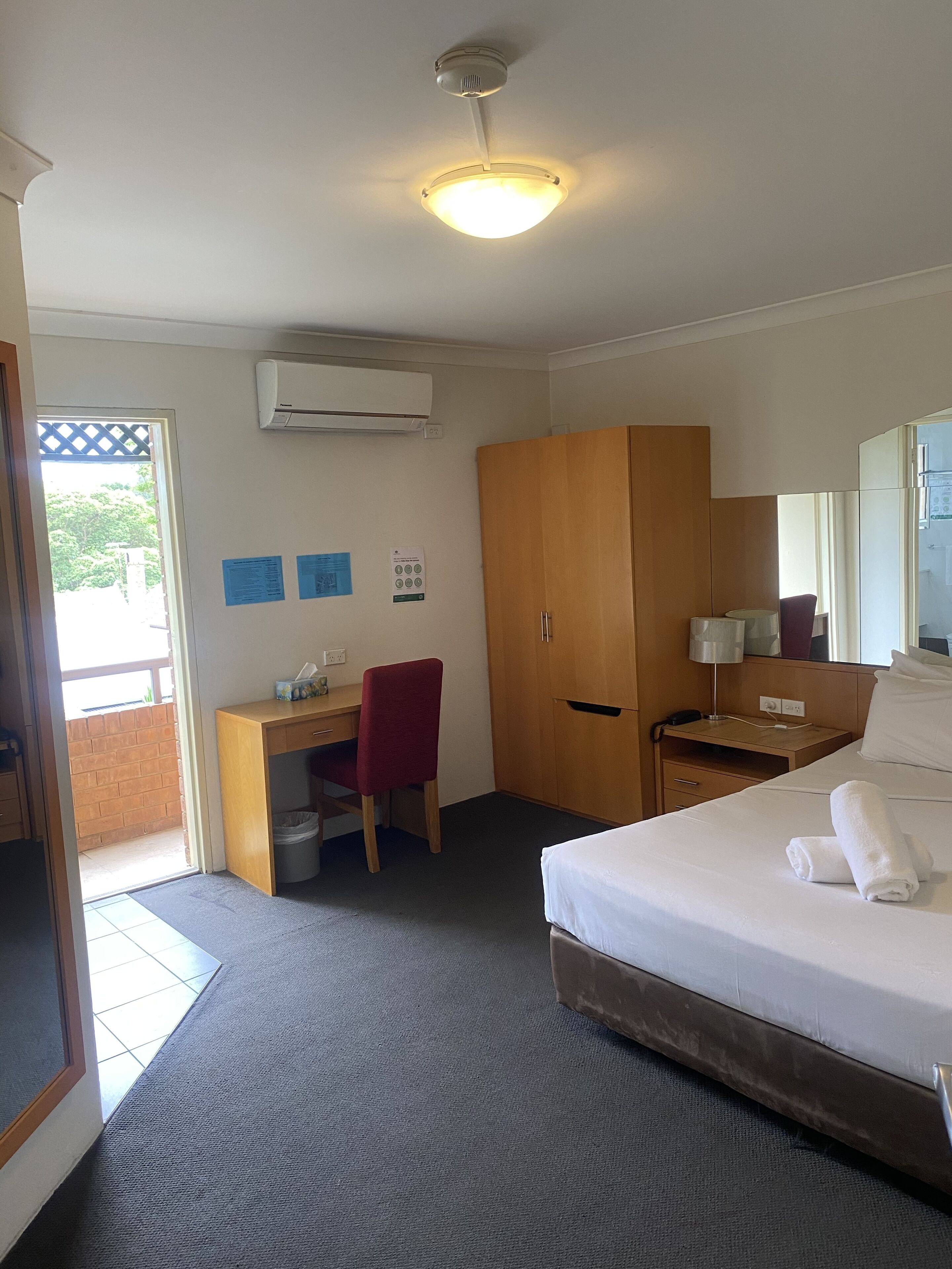 A hotel room with a bed, desk, chair, wardrobe, and an air conditioning unit.