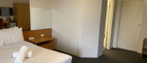A hotel room with a bed, a bedside table, a mirror, a wardrobe, a desk with a chair, and a door.