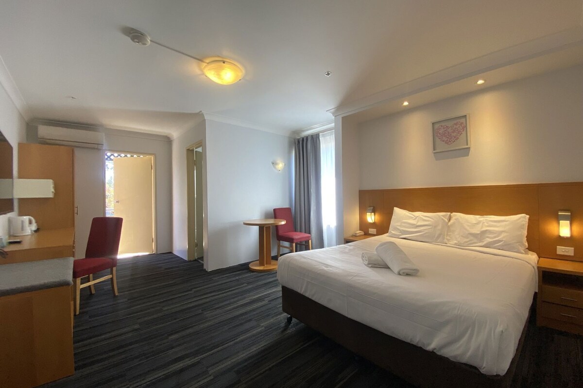 A hotel room with a bed, a desk, a chair, a small table, and a wall-mounted light.
