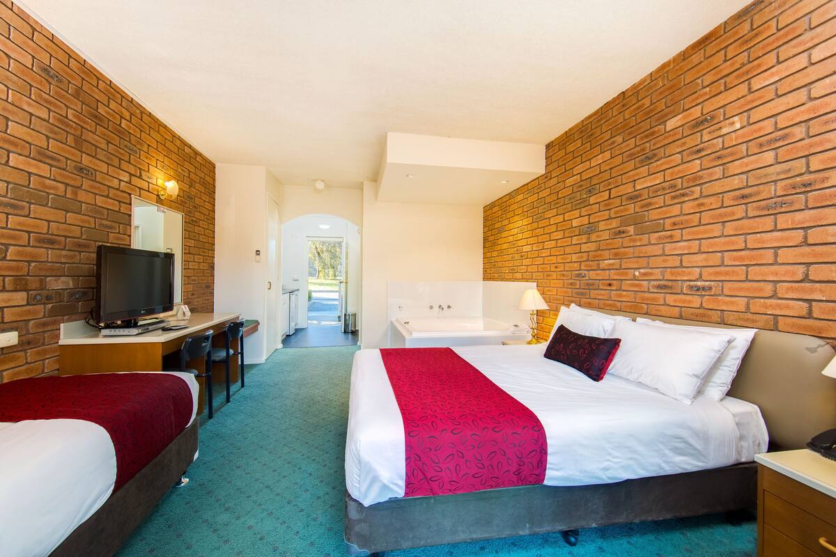 A hotel room with a brick wall, a bed with white bedding and a red accent, a desk with a TV, and a bathroom with a bathtub and a mirror.