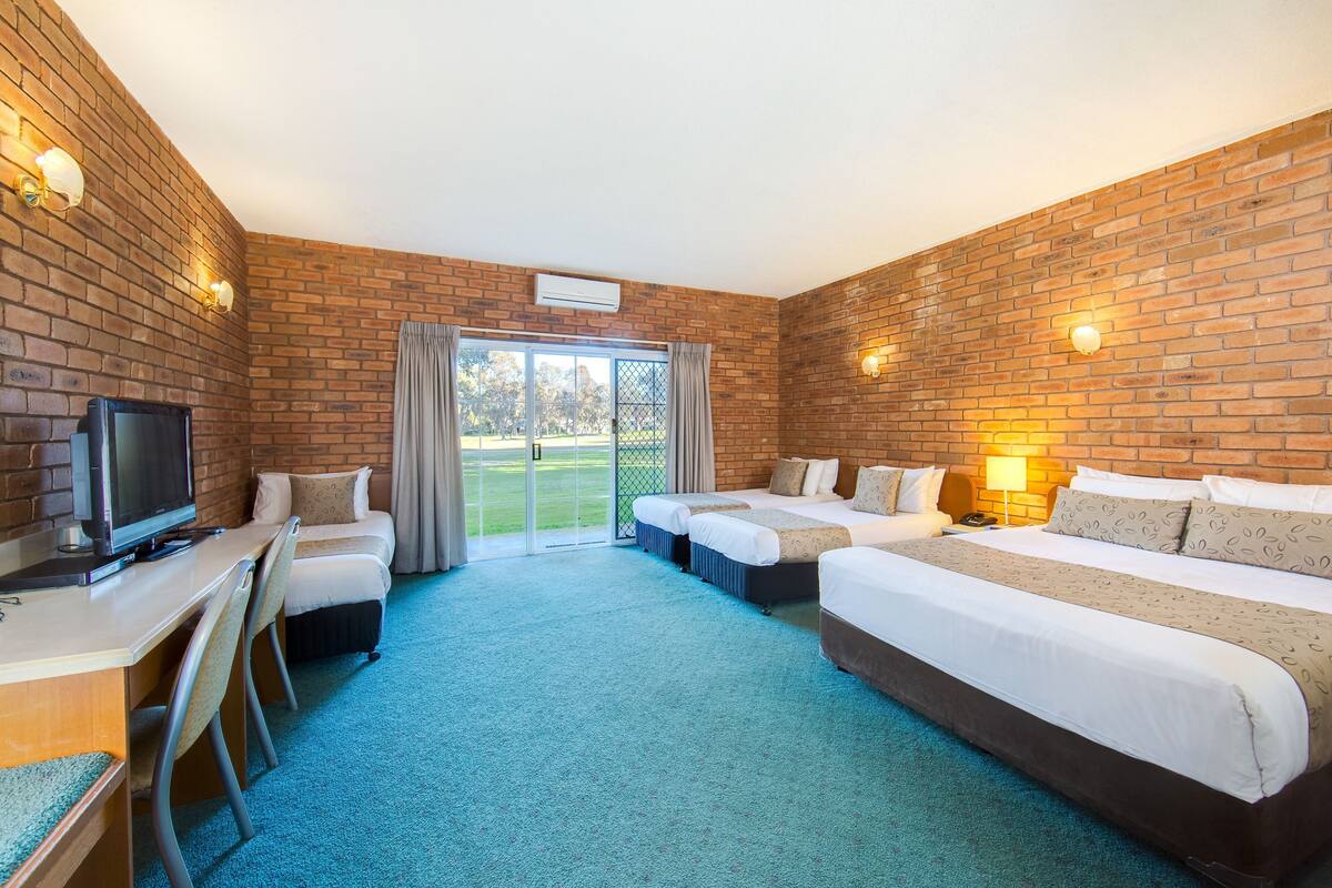 A hotel room with a brick wall, a TV, two beds, a desk, and a view through a glass door.
