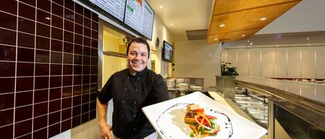 Un chef tenant une assiette de nourriture dans une cuisine moderne aux murs carrelés et équipée d’appareils électroménagers en acier inoxydable.