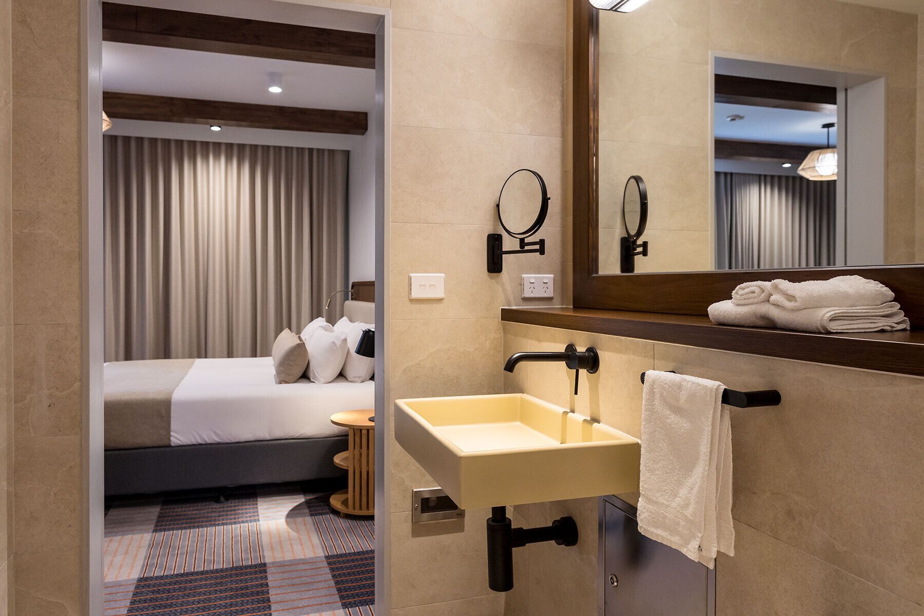 A modern hotel bathroom with a large mirror, a sink, and a bed visible in the background.