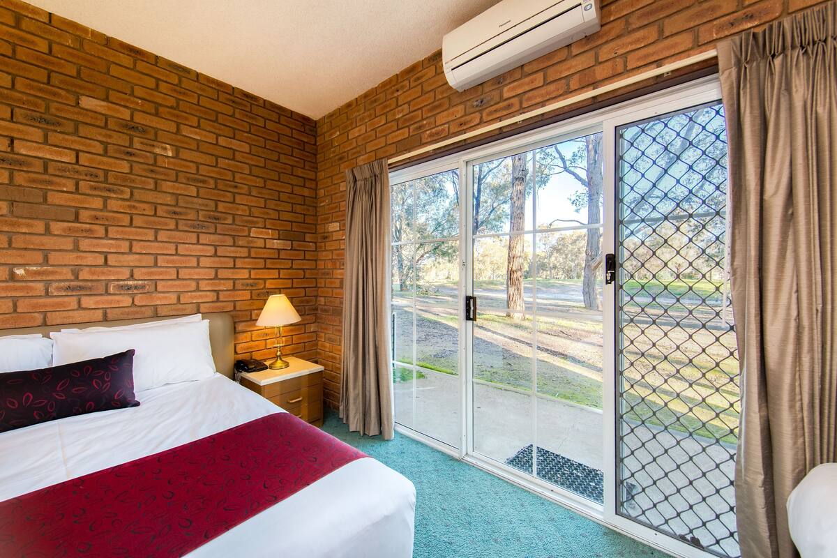 A hotel room with a brick wall, a large bed with white bedding, a nightstand with a lamp, and a sliding glass door leading to an outdoor area.