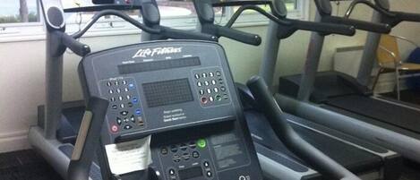 A treadmill with a digital display and control panel, positioned in a room with windows and a view of trees outside.