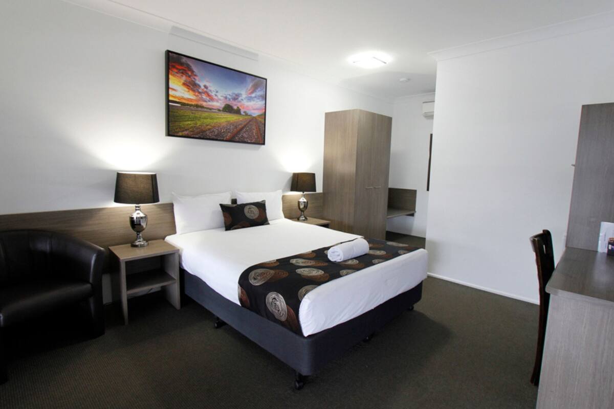 Habitación de hotel con cama, dos mesitas de noche, escritorio, silla, armario y un cuadro en la pared.
