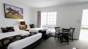 A hotel room with two beds, a dining table, and a chair. There is a window with curtains, a painting on the wall, and a lamp on the nightstand. - Takalvan Motel (Bundaberg West)