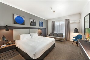 A modern hotel room with a large bed, a desk, a chair, and a sofa.