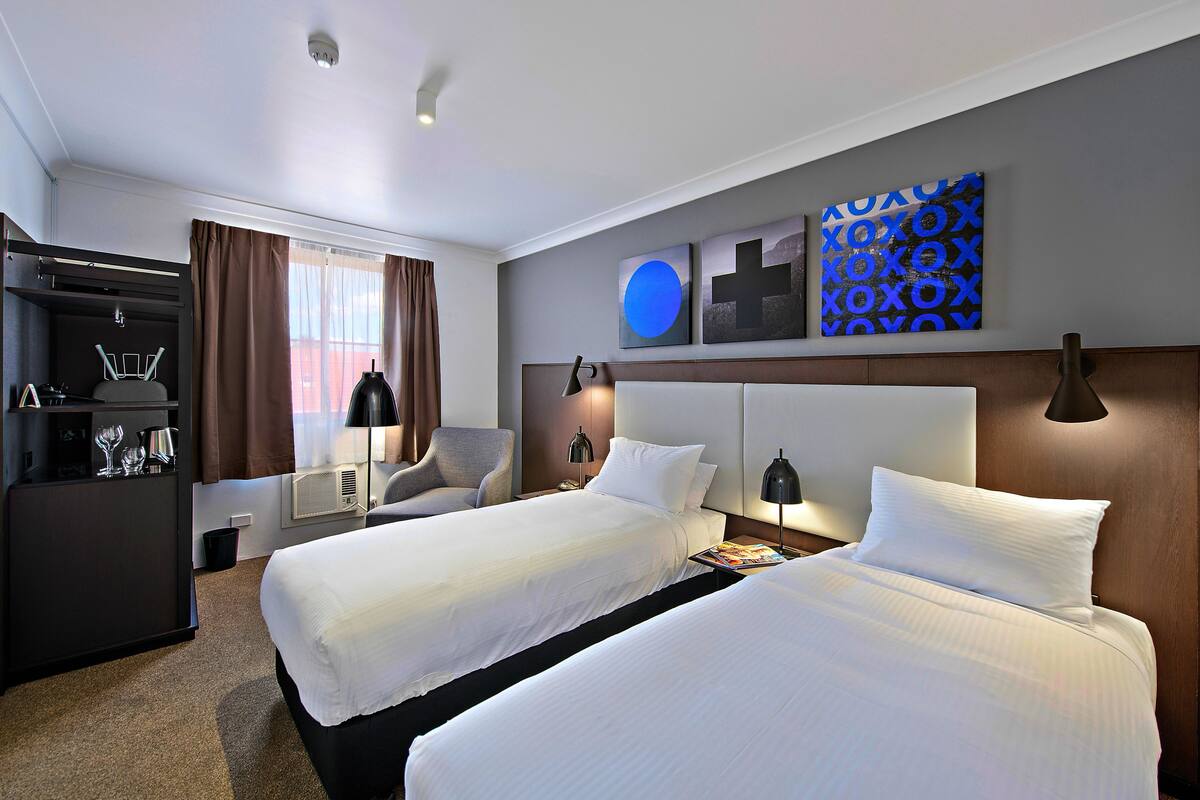 Une chambre d’hôtel avec deux lits, un bureau avec une chaise, un minibar et des œuvres d’art accrochées au mur.