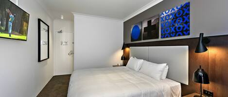 A modern hotel room with a large bed, a flat-screen TV mounted on the wall, a wooden headboard, and a nightstand with a lamp.