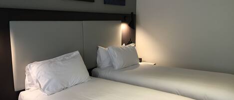 Habitación de hotel con dos camas individuales, ropa de cama blanca, cabecero, lámparas de noche y una obra de arte colgada en la pared.