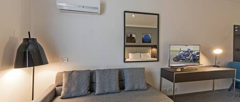 Una habitación moderna con un sofá gris, un escritorio con computadora y lámpara, un espejo y aire acondicionado.
