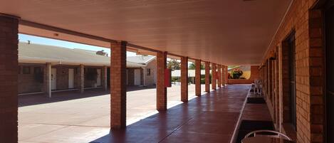 A covered outdoor area with brick pillars and a tiled floor, leading to a building with multiple doors.