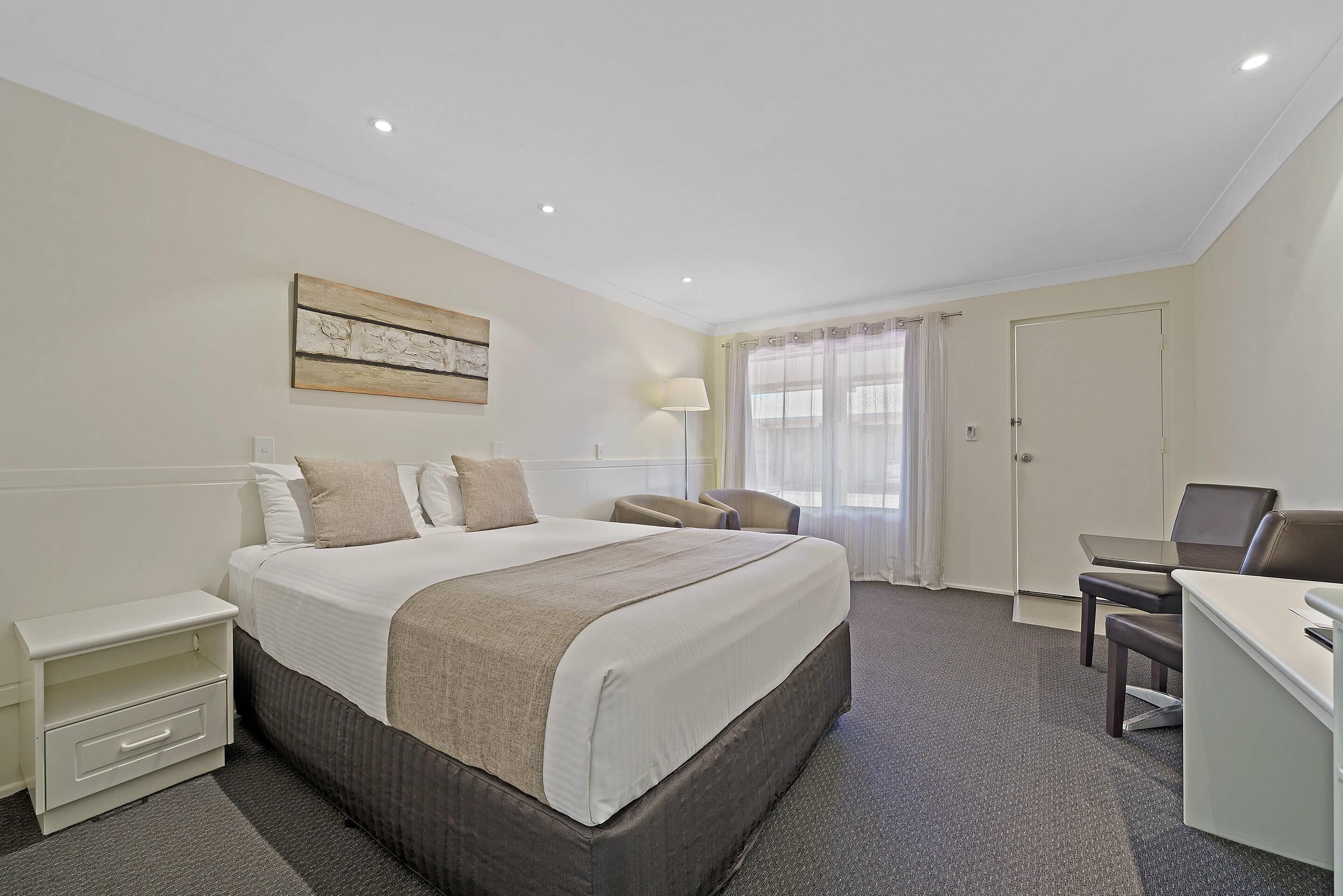 A hotel room with a large bed, a desk, a chair, a nightstand, and a lamp.