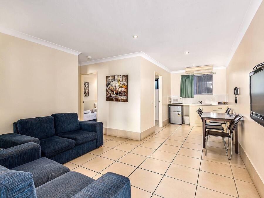 A modern living room with a sofa, a dining table with chairs, a kitchen area, and a flat-screen TV mounted on the wall.