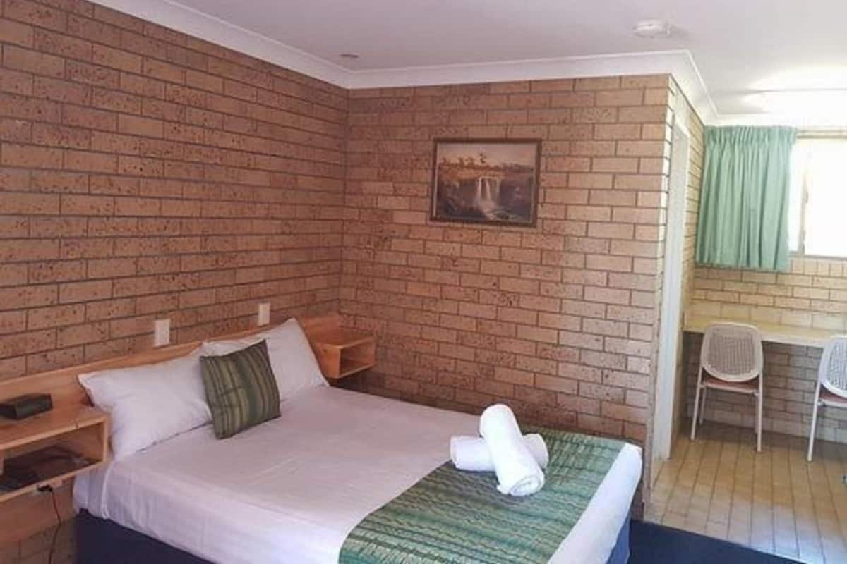 A single bed with a green blanket, a wooden nightstand, a painting on the wall, and a window with curtains.