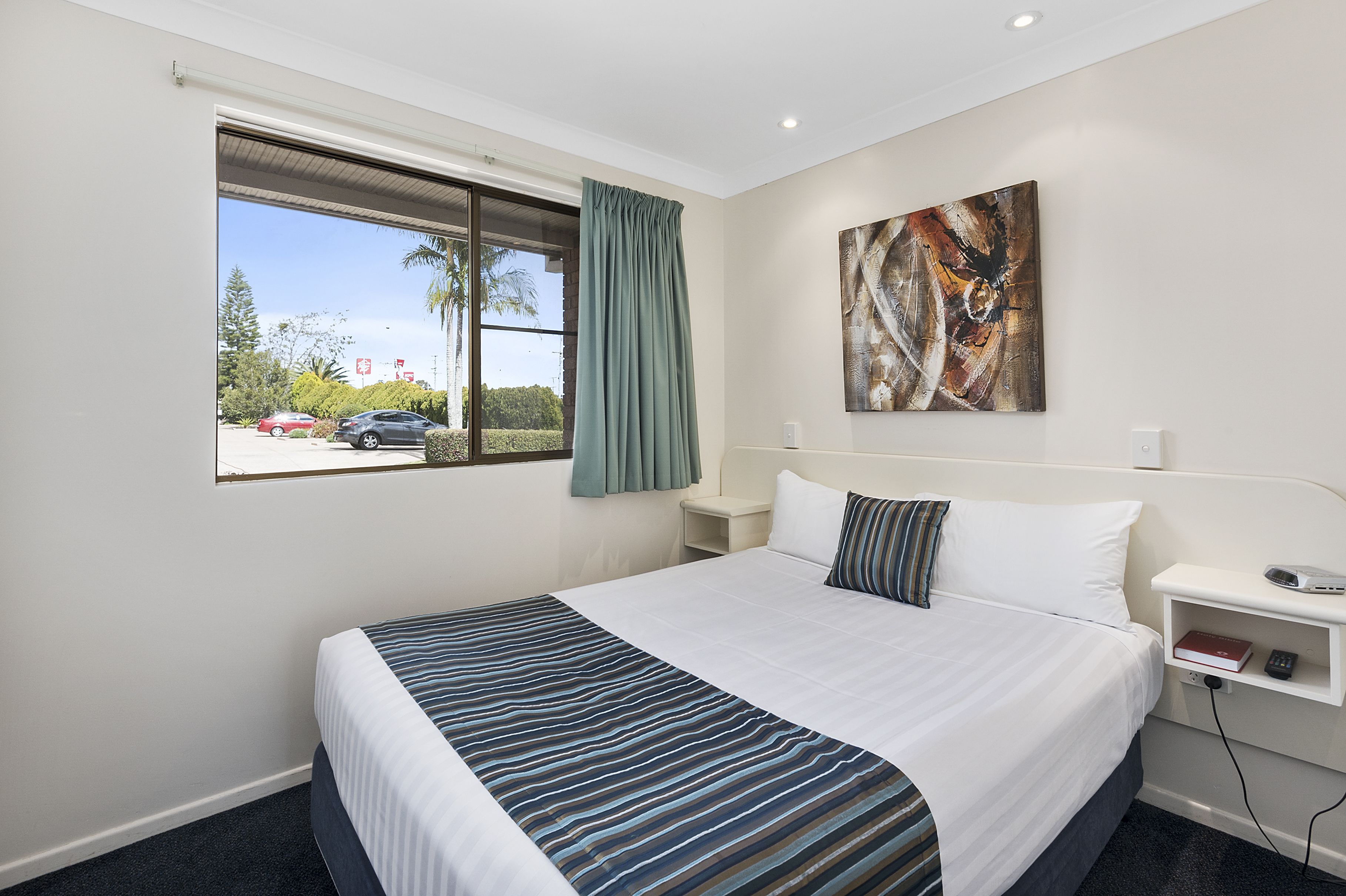 Habitación de hotel con una cama grande, una mesita de noche con lámpara, una ventana con vista a un estacionamiento y un cuadro en la pared.