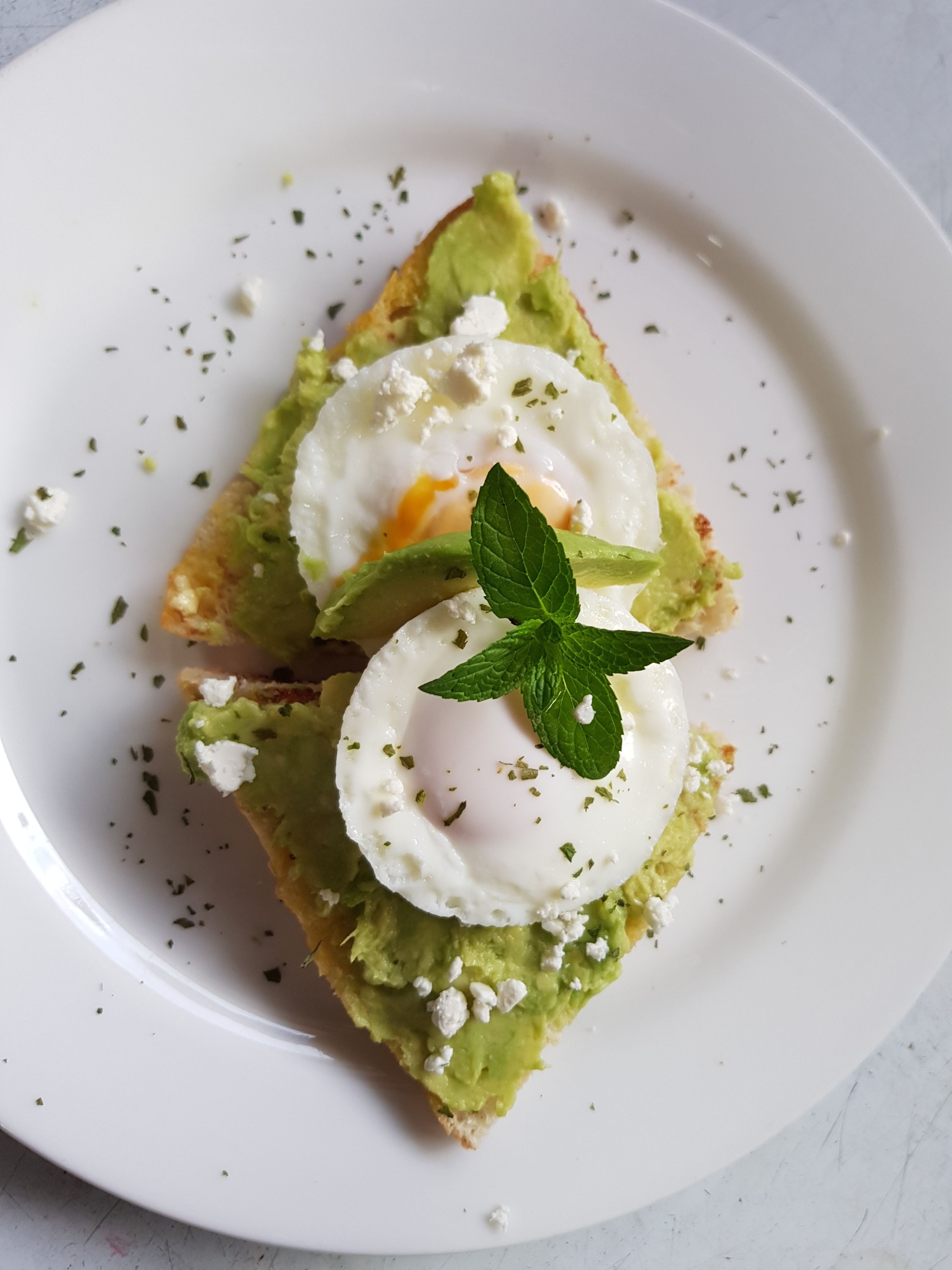 Une assiette avec une tranche de pain grillé garnie d’avocat, d’un œuf poché et de fromage feta, le tout décoré d’une feuille de menthe.