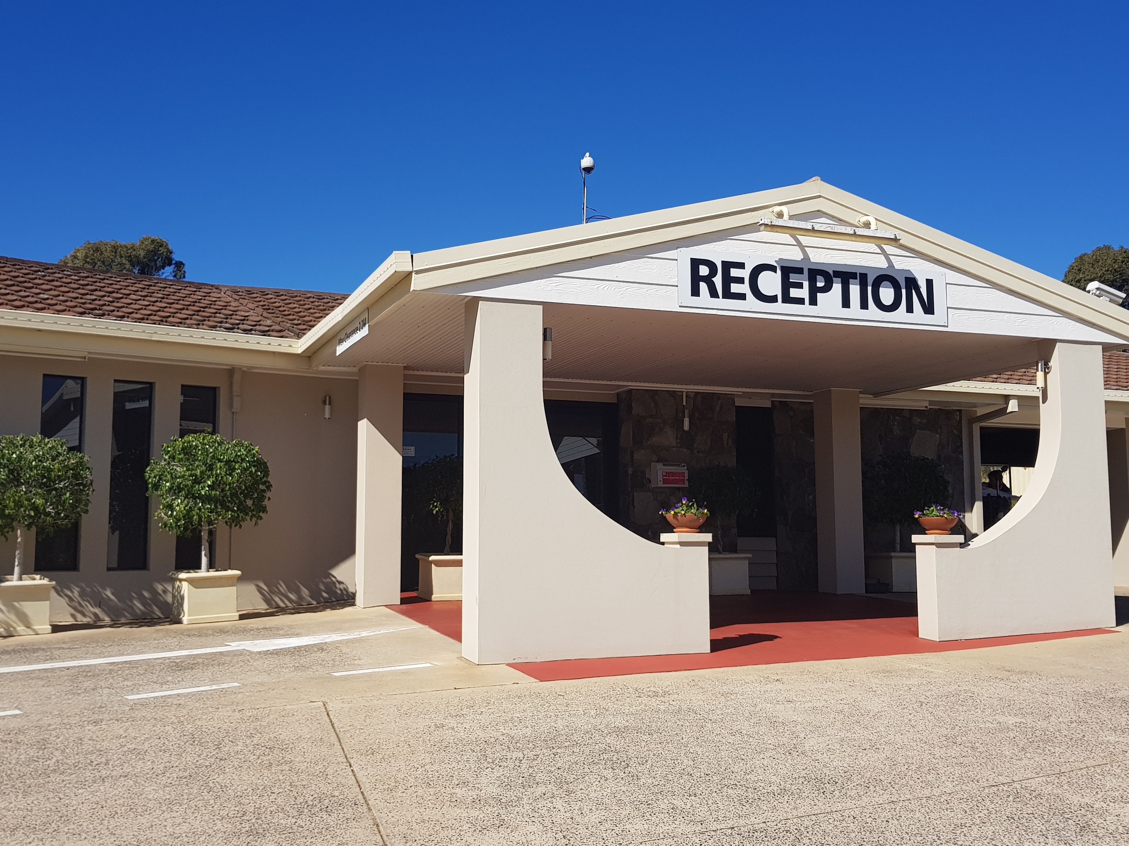 A building with a reception area, featuring a tiled roof, a signboard, and potted plants.