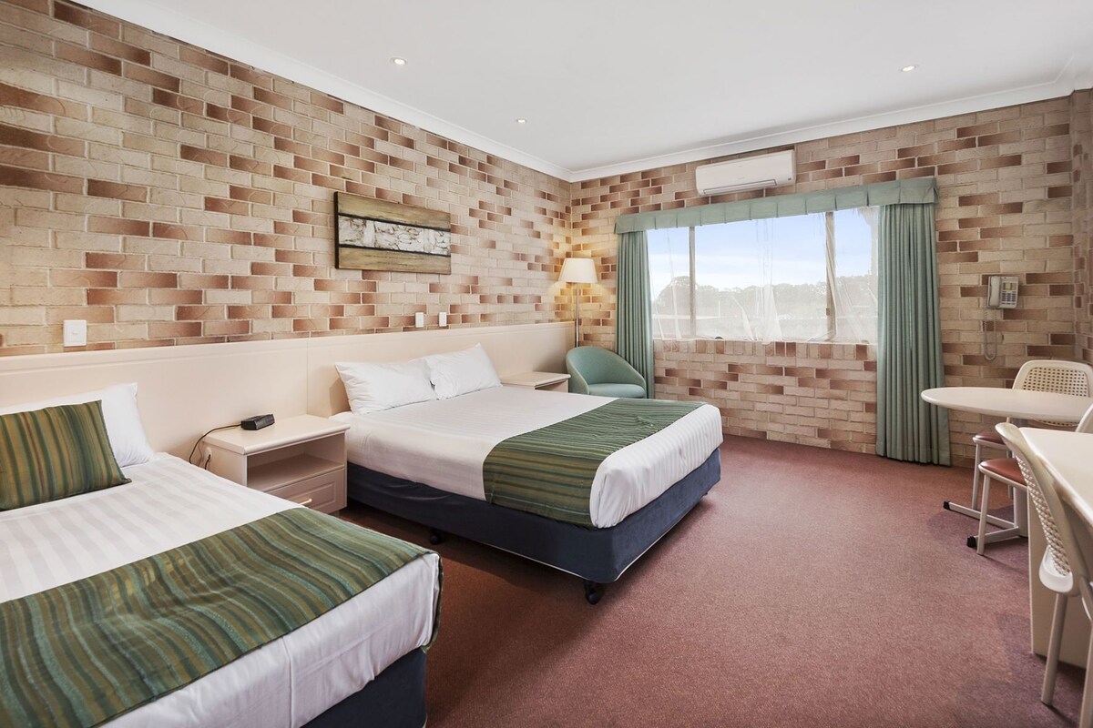 A hotel room with two beds, a desk, a chair, a window with curtains, and a brick wall.
