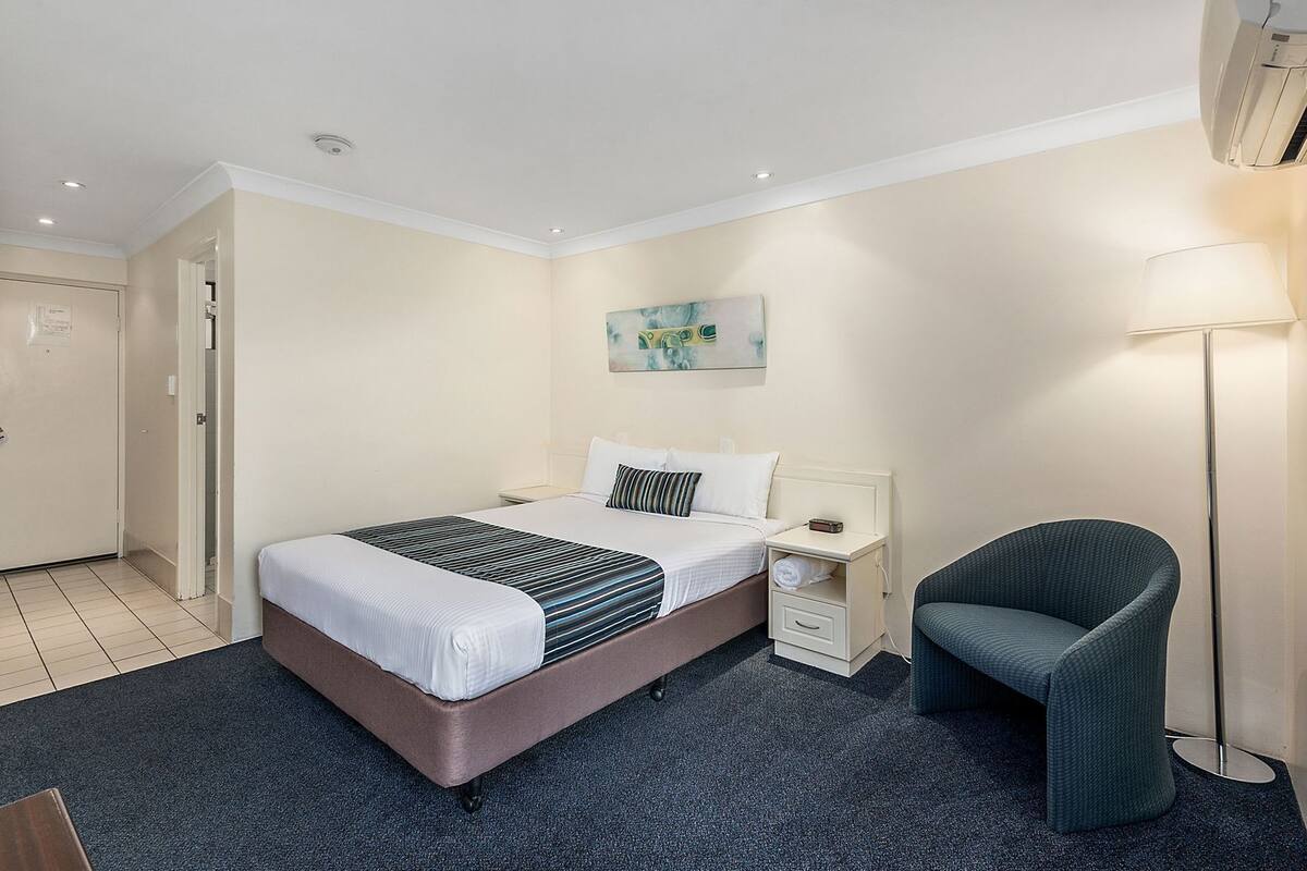 A hotel room with a bed, bedside table, chair, and a painting on the wall.
