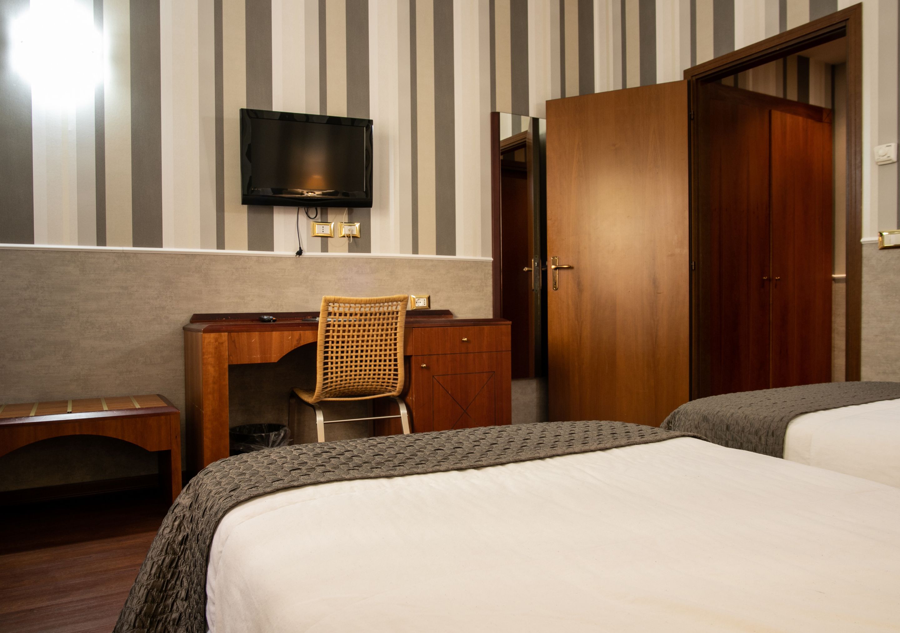 A hotel room with a bed, a desk with a chair, a television mounted on the wall, and a wardrobe.