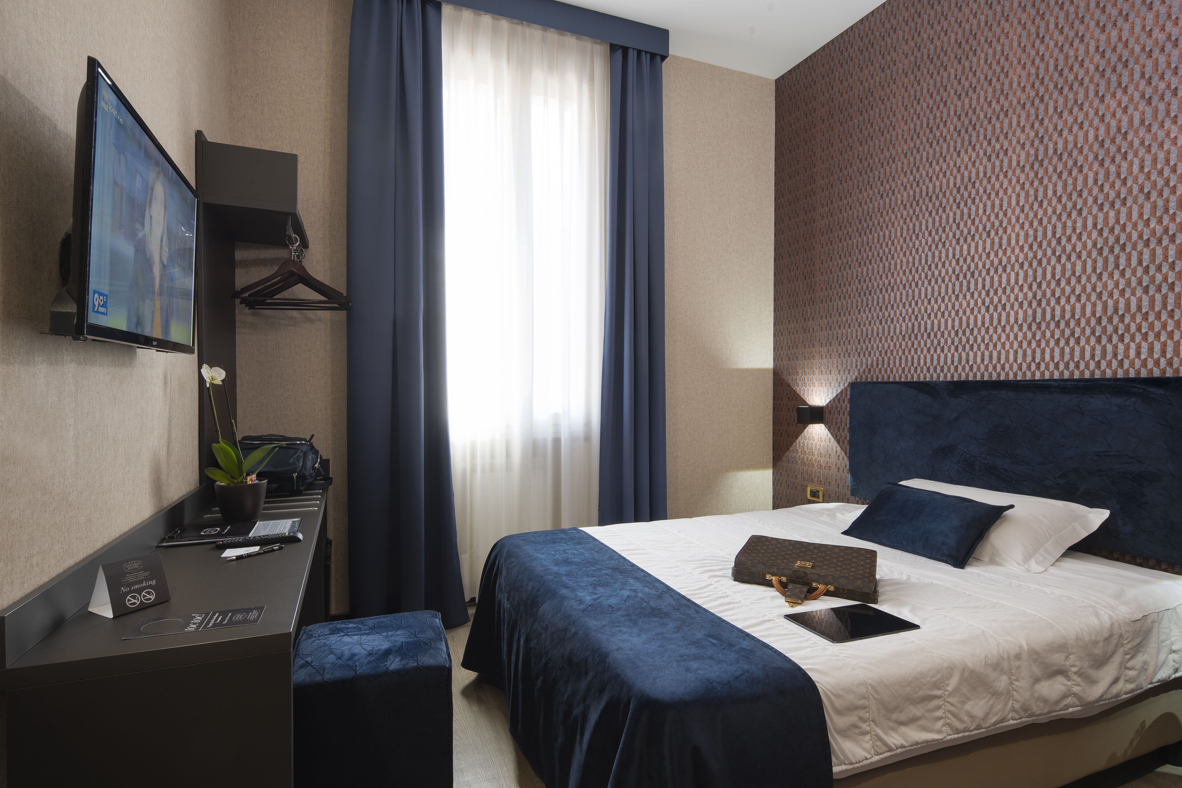 Ein Hotelzimmer mit Bett, Schreibtisch mit Computer, Fernseher und einem Fenster mit Vorhängen.