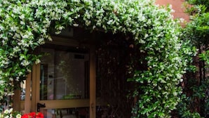 A garden with a wooden pergola covered in climbing plants, a glass door entrance, and a planter with red flowers. - Hotel Astoria (Bologna)