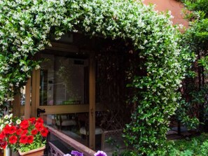 A garden with a wooden pergola covered in climbing plants, a glass door entrance, and a planter with red flowers. - Hotel Astoria (Bologna)