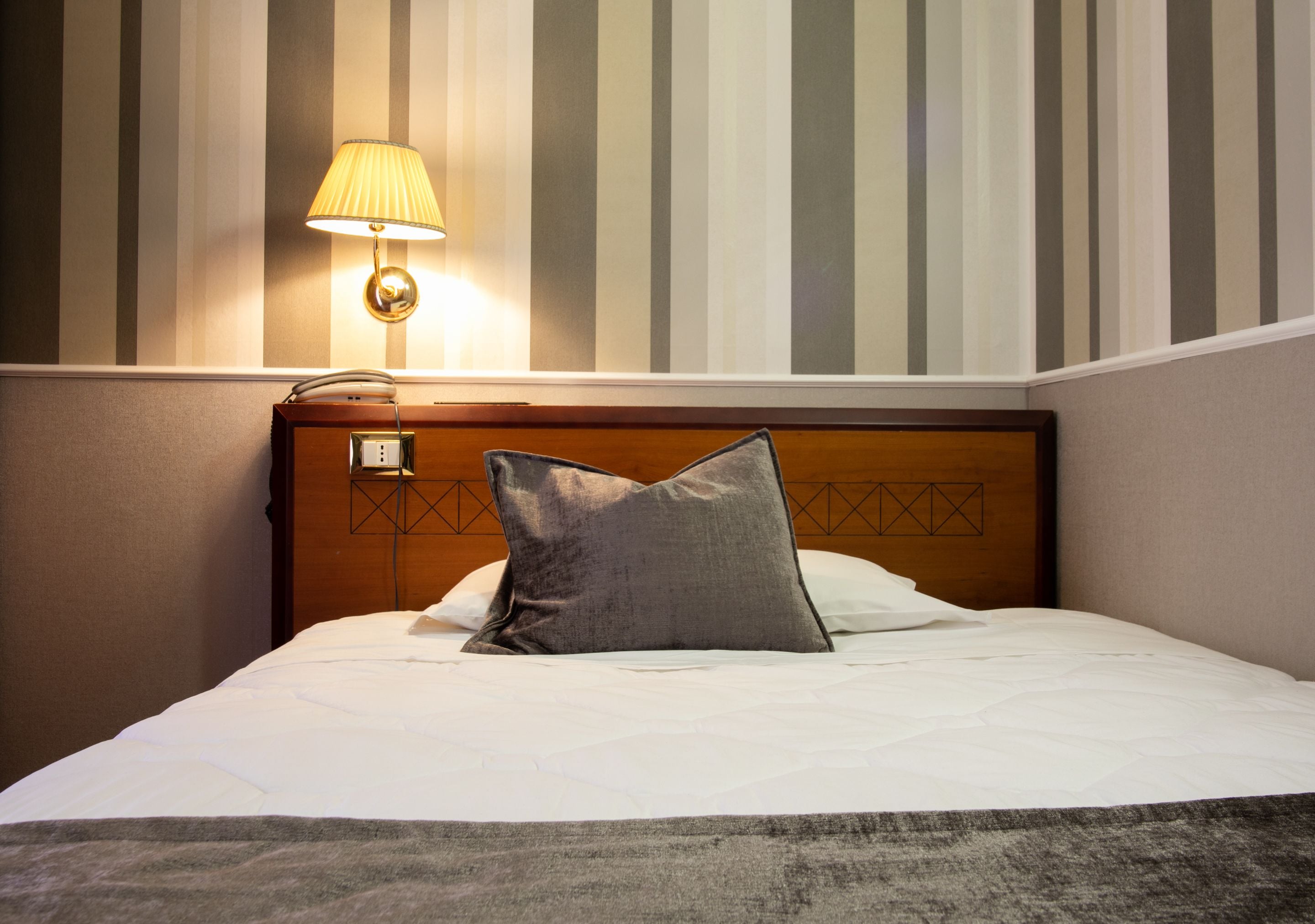 Ein Hotelzimmer mit einem ordentlich bezogenem Bett, einer Wandleuchte und rauen Tapeten.