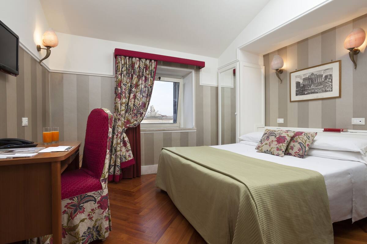A hotel room with a bed, a desk with a chair, a window with curtains, and a wall-mounted picture.