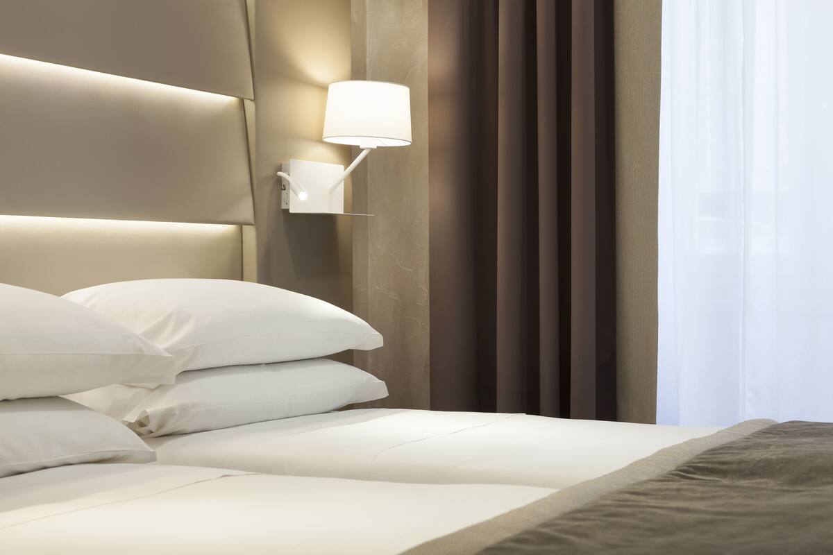 A neatly made bed with white bedding and a headboard, a wall-mounted lamp, and a window with curtains.