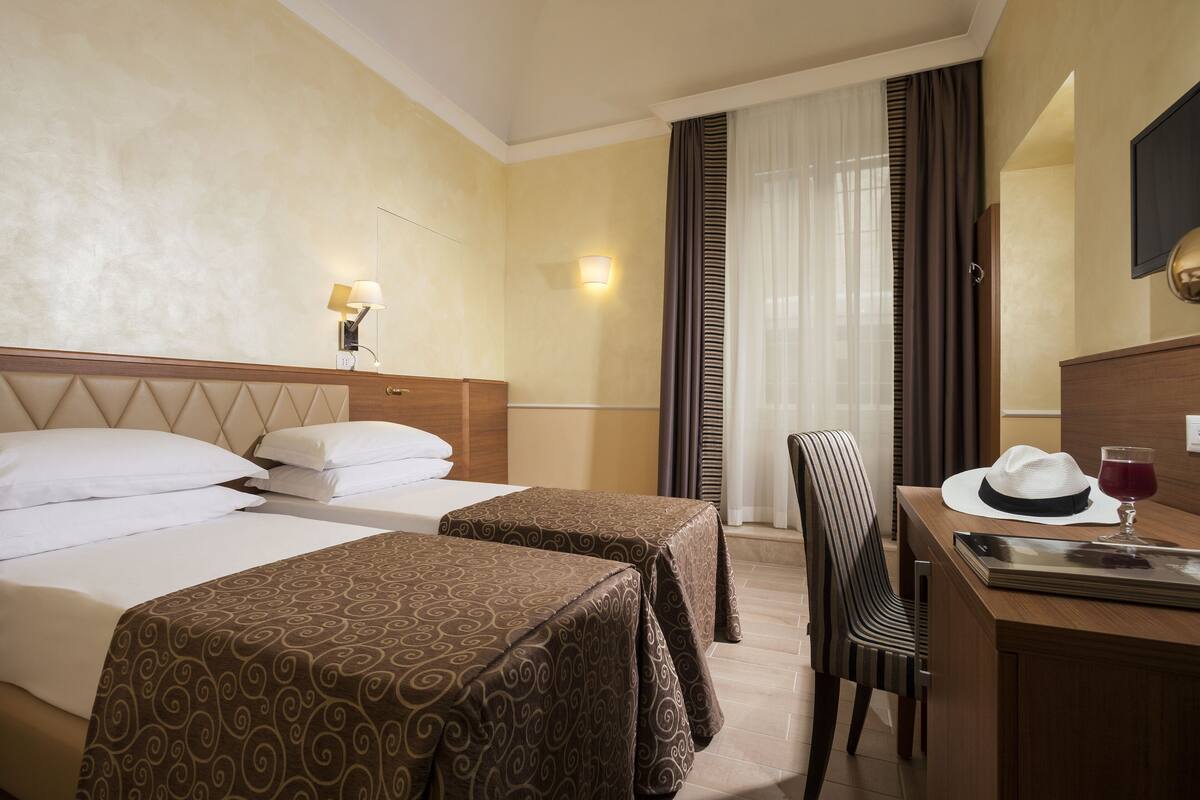 A hotel room with two beds, a desk, a chair, a lamp, and a window with curtains.