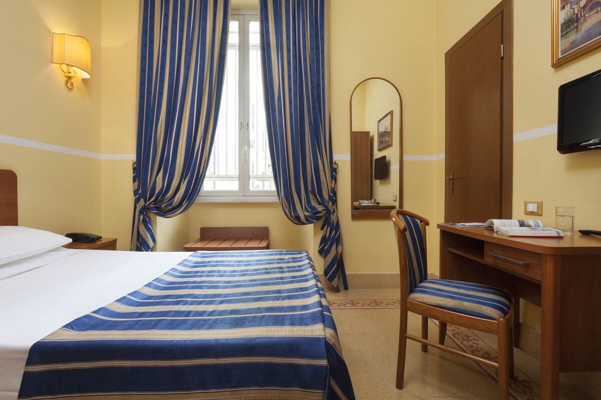 A hotel room with a bed, a desk, a chair, a mirror, and curtains.