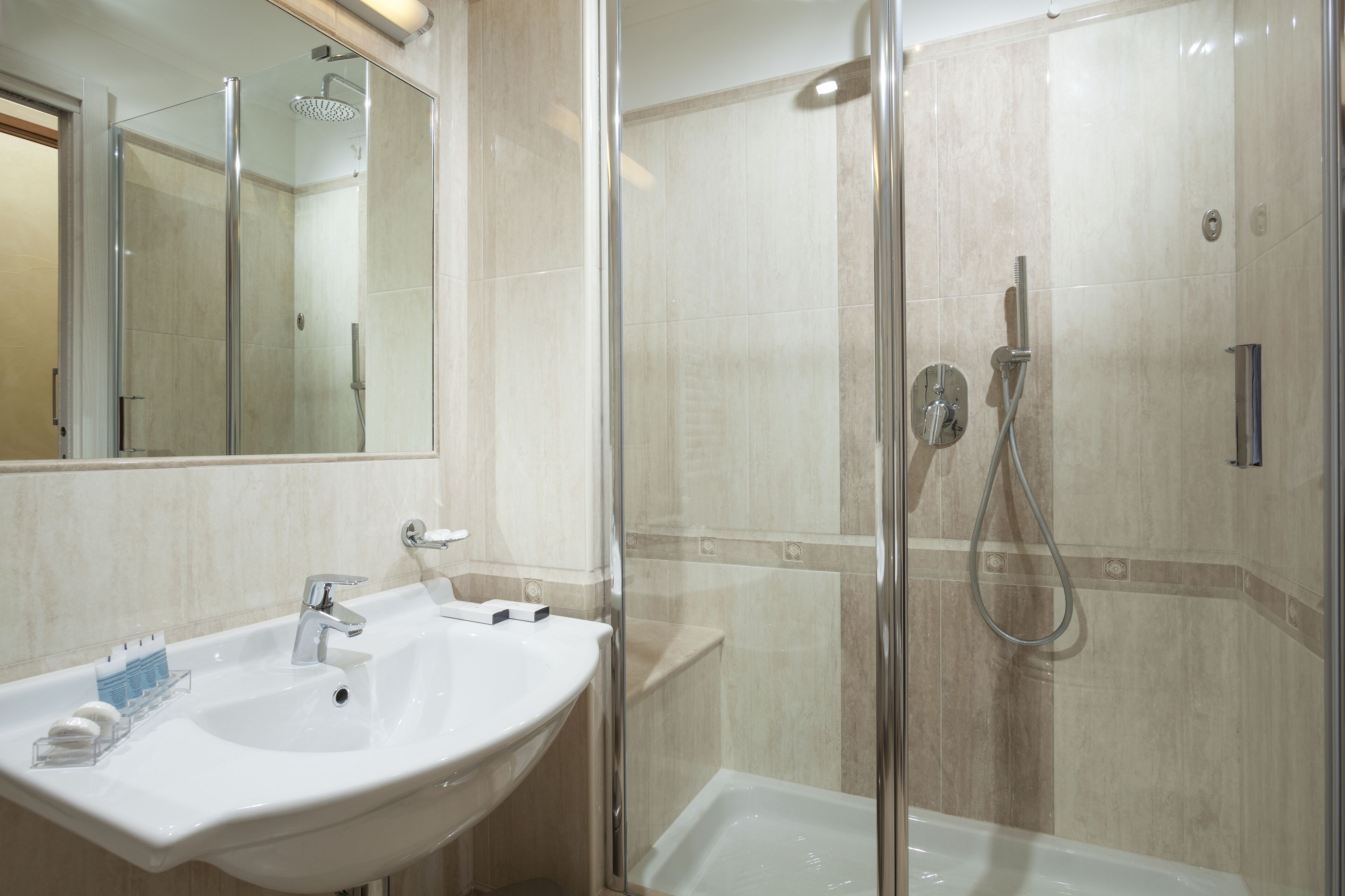Un bagno moderno con doccia vetrata, lavandino bianco con specchio e pareti rivestite di piastrelle color crema.