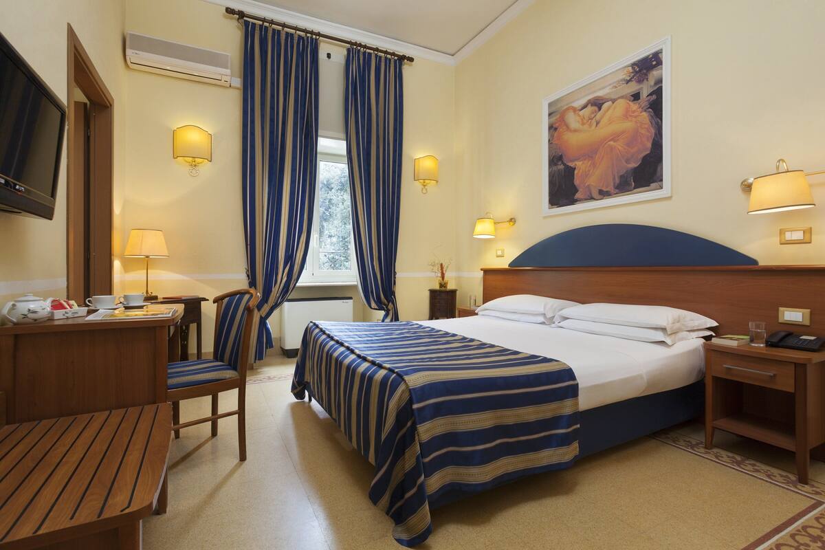 A hotel room with a bed, a desk, a chair, a TV, and a painting on the wall.