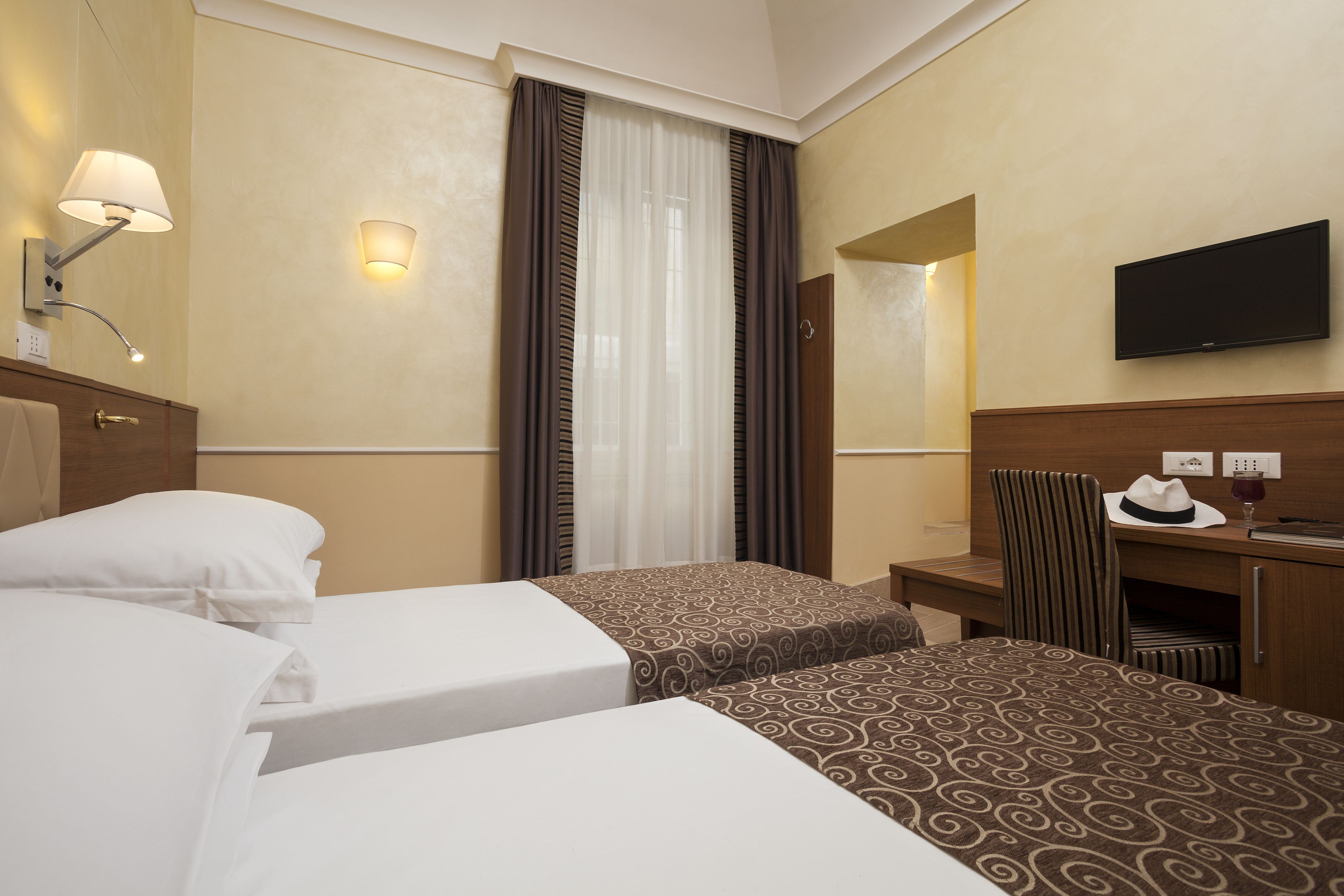 A hotel room with two beds, a desk, a chair, a television, and a window with curtains.