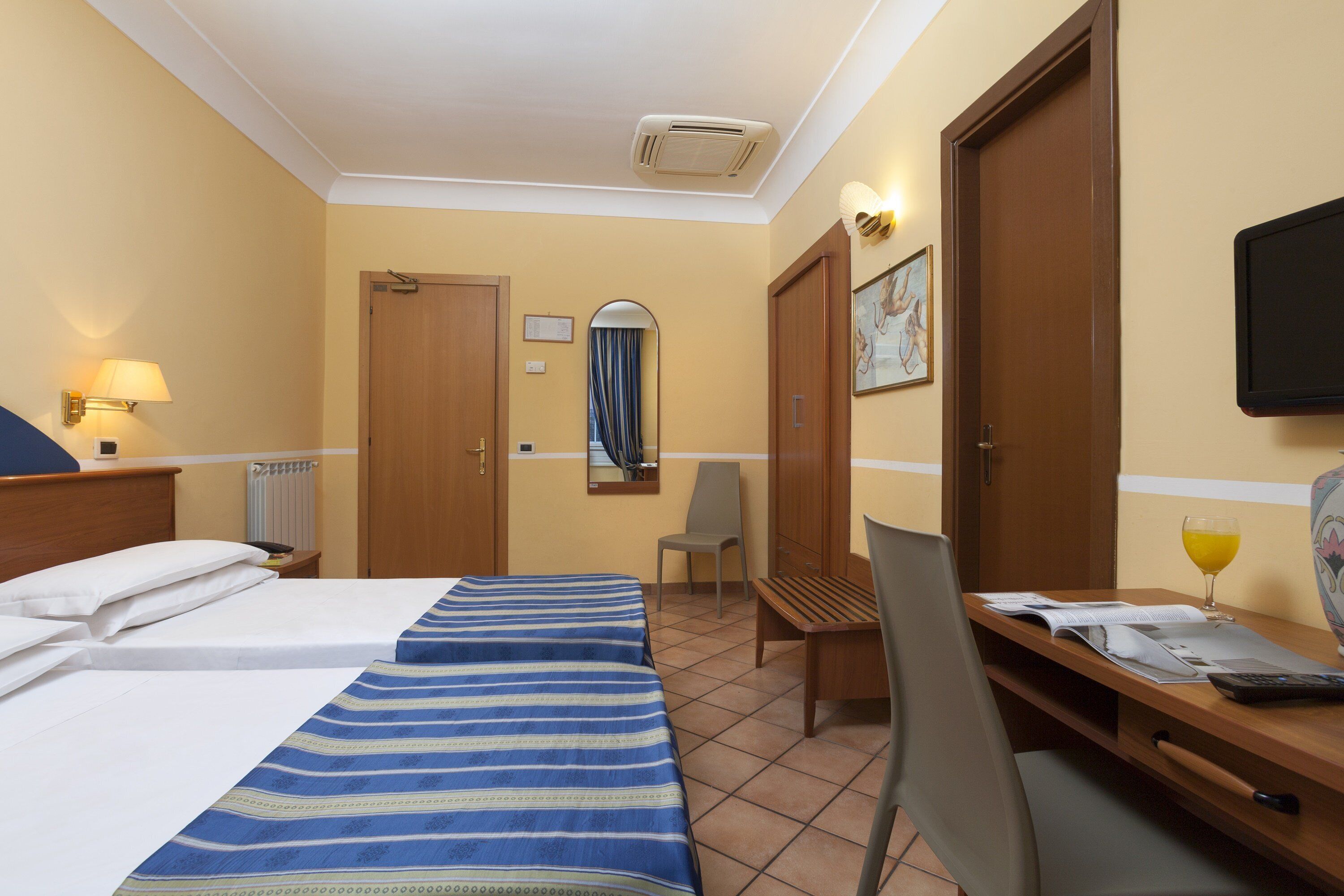 Habitació d'hotel amb dos llits, una taula de treball, una cadira, un televisor i una finestra amb cortines.