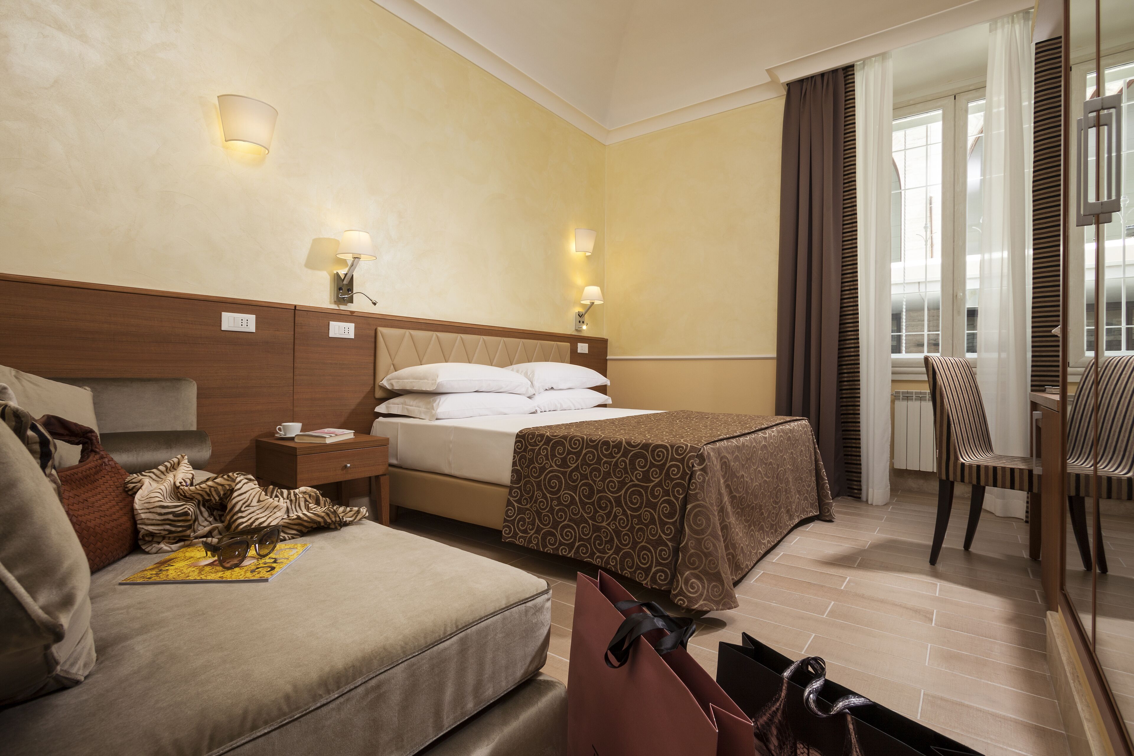 Habitación de hotel con cama, sofá, mesita de noche, lámpara, silla y vista al exterior.