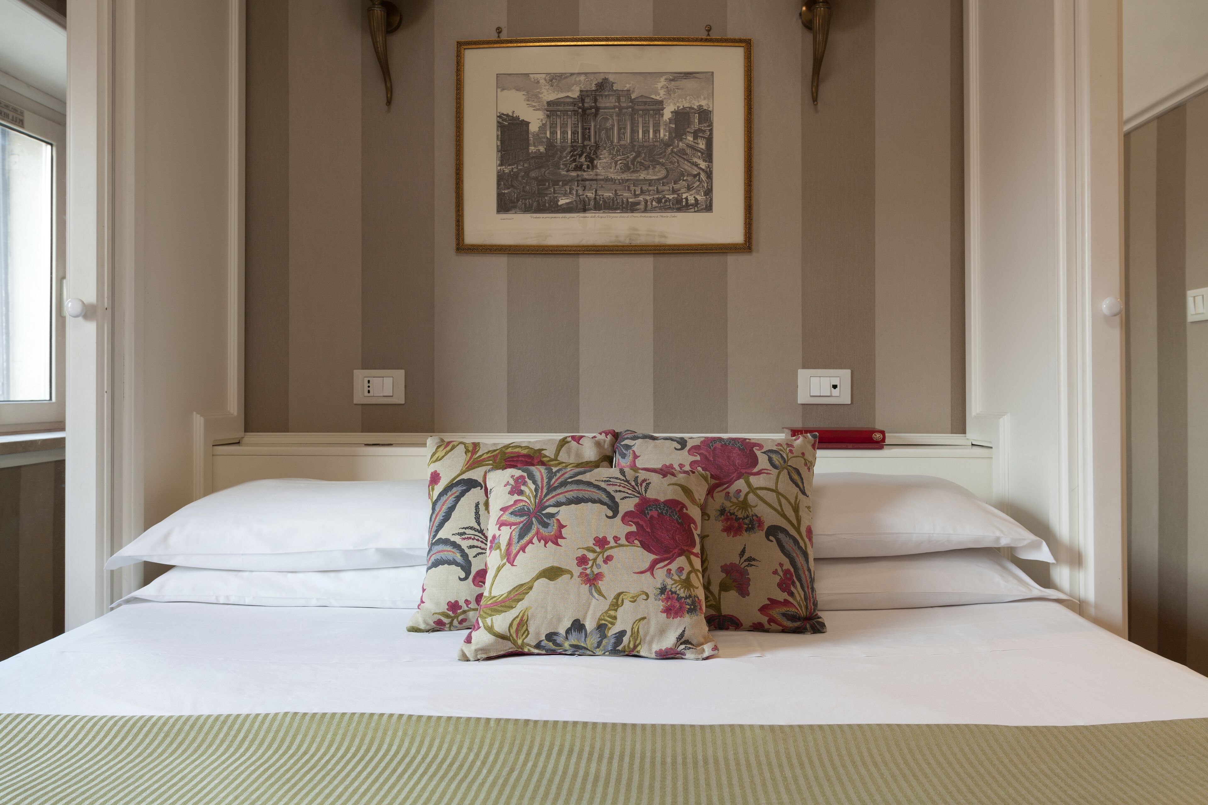 Une chambre d’hôtel avec un lit, des oreillers à motifs floraux, un tableau encadré au mur et une fenêtre.