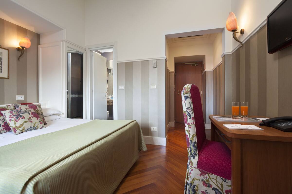 A hotel room with a bed, a desk with a telephone and glasses of orange juice, and a television on the wall.