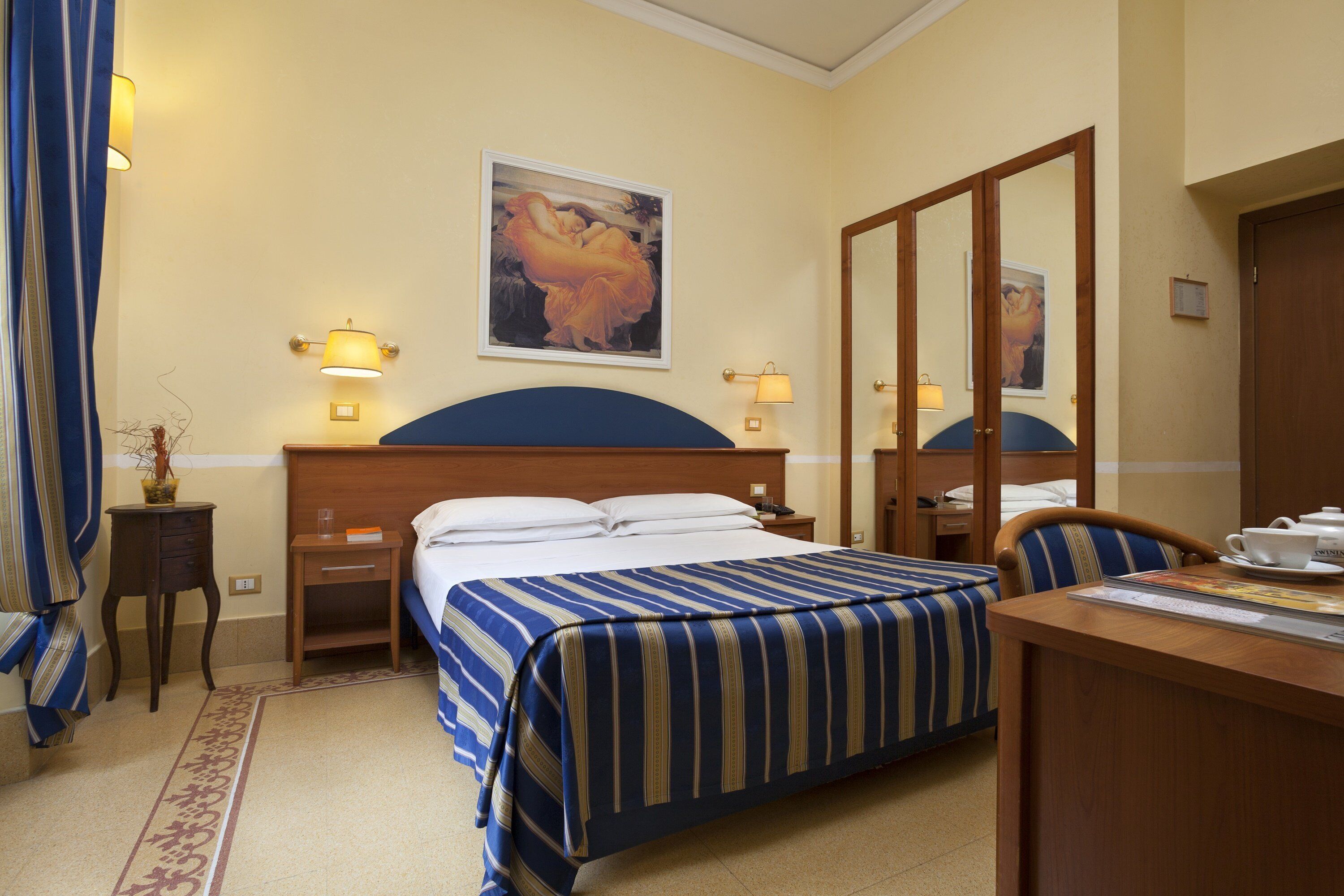 A hotel room with a large bed, a desk with a chair, a nightstand, and a wall-mounted lamp.