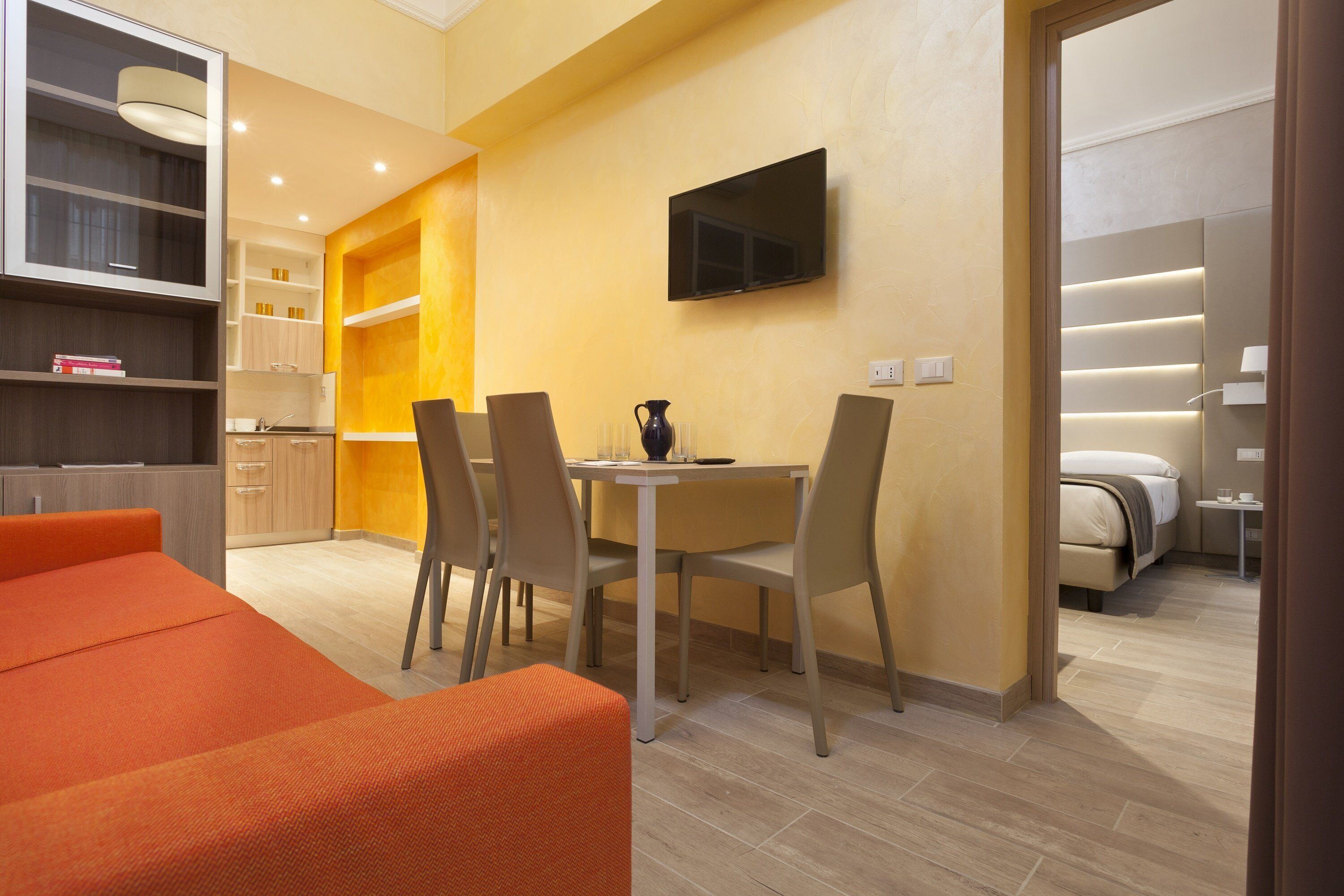 A modern hotel room with a dining area, a TV mounted on the wall, and a bed with a bedside table.