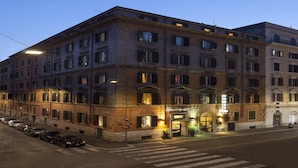 Un hotel de varios pisos con un estilo arquitectónico clásico, que presenta una entrada central con dosel, múltiples ventanas y un área de estacionamiento al frente.