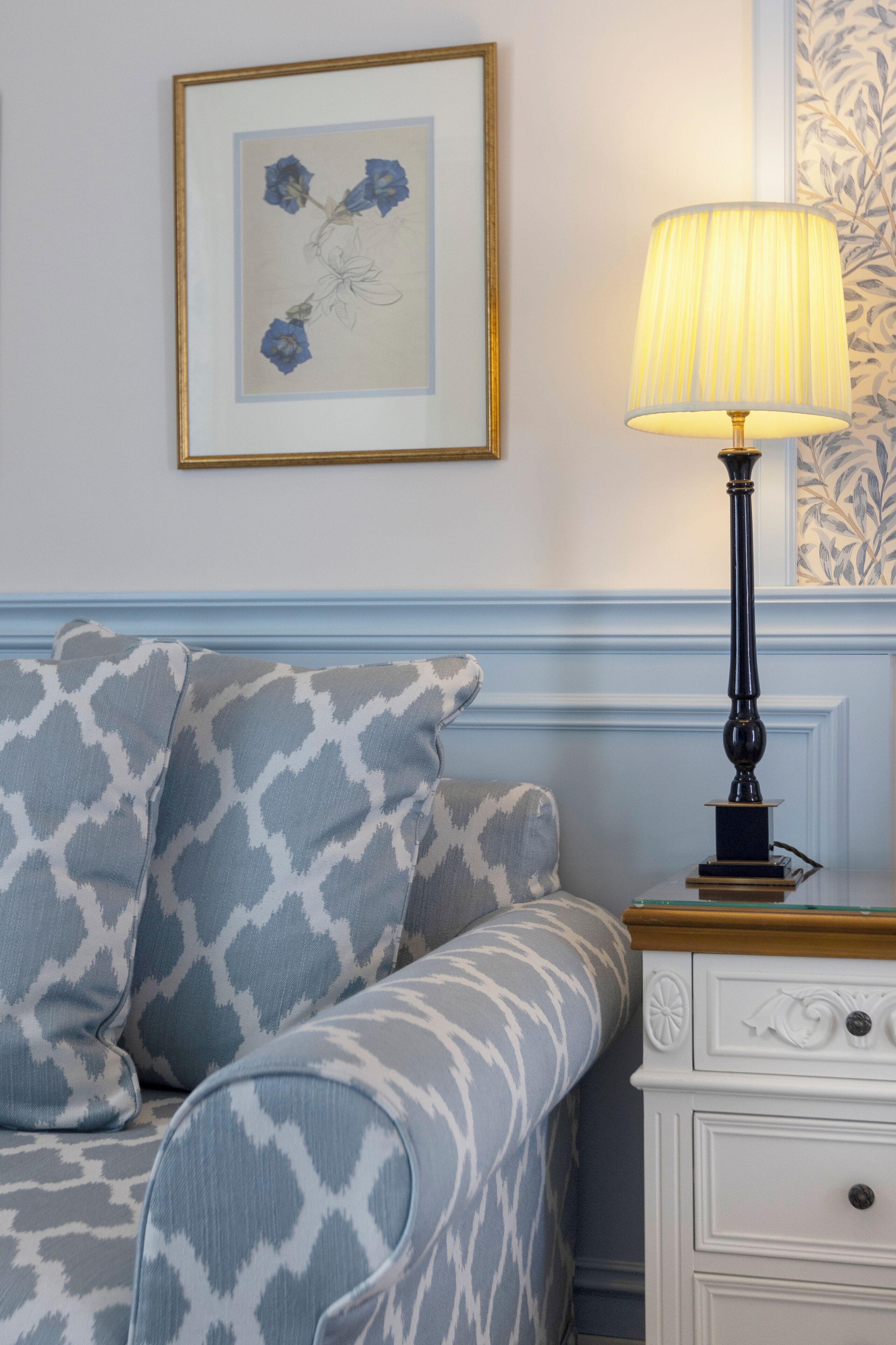 A room with a patterned sofa, a framed floral artwork on the wall, a lamp on a nightstand, and a patterned wallpaper.