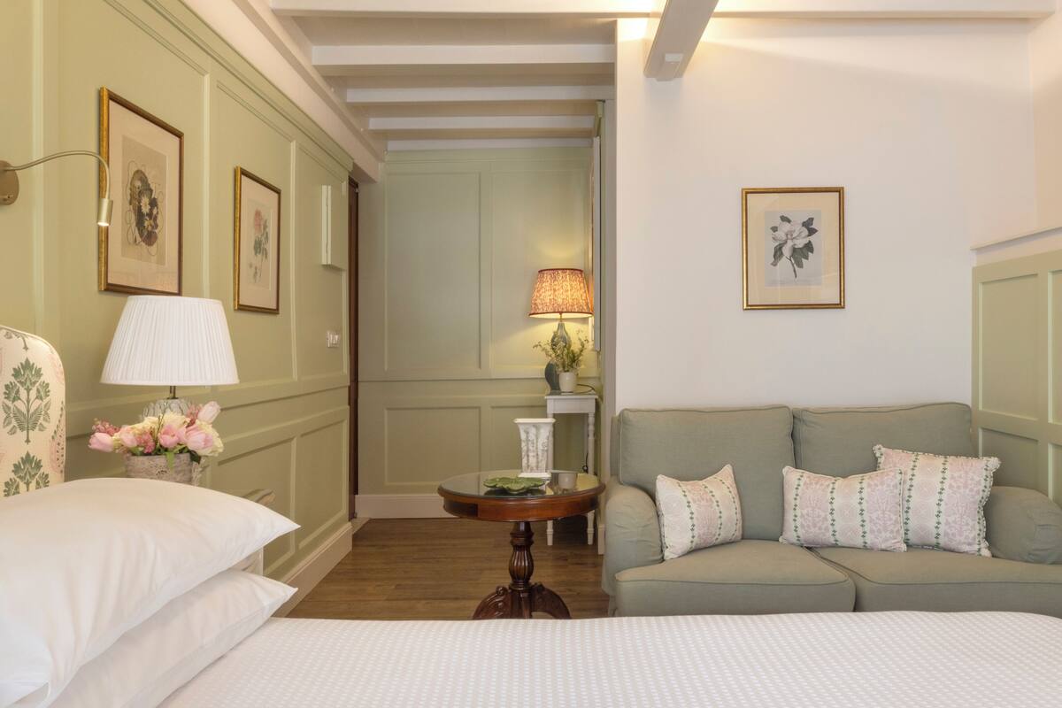 A hotel room with a bed, a sofa, a small table with a lamp, and framed pictures on the walls.