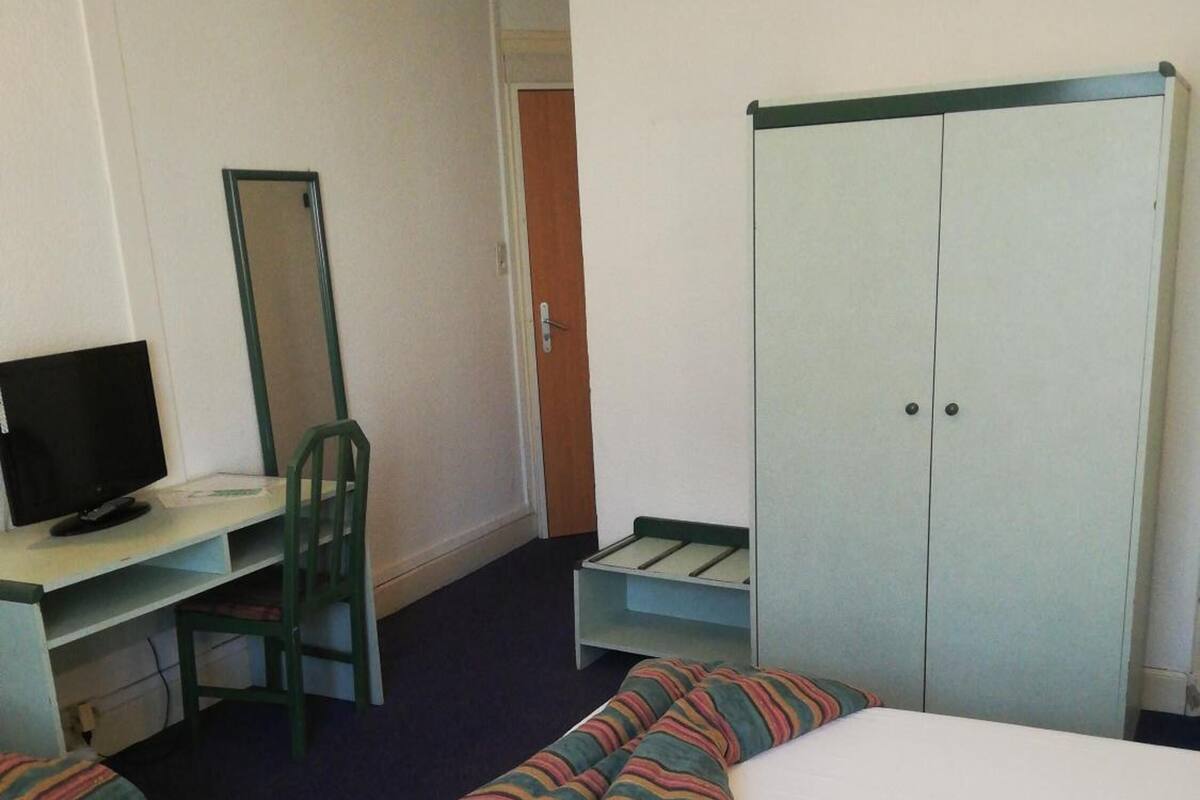A hotel room with a bed, a desk with a computer, a chair, a wardrobe, and a mirror.