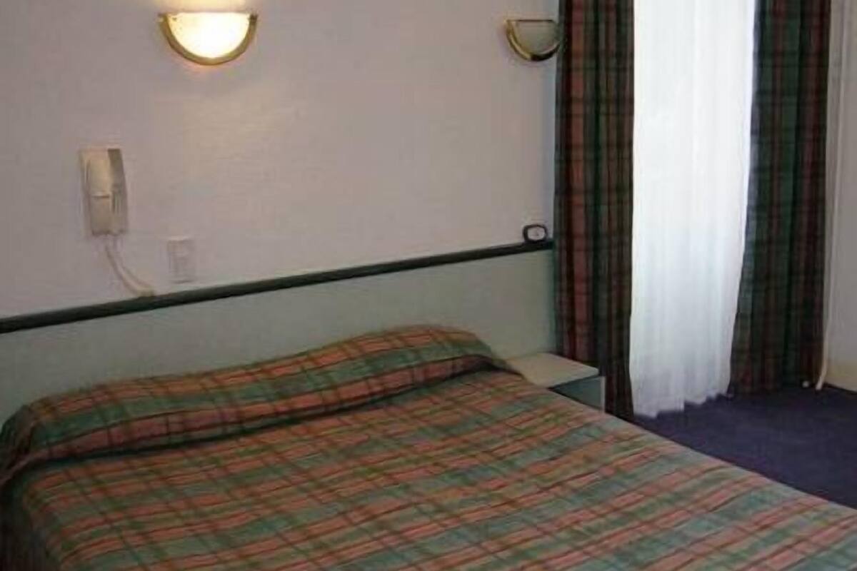 A single bed with a plaid bedspread, a wall-mounted light, and a window with curtains.