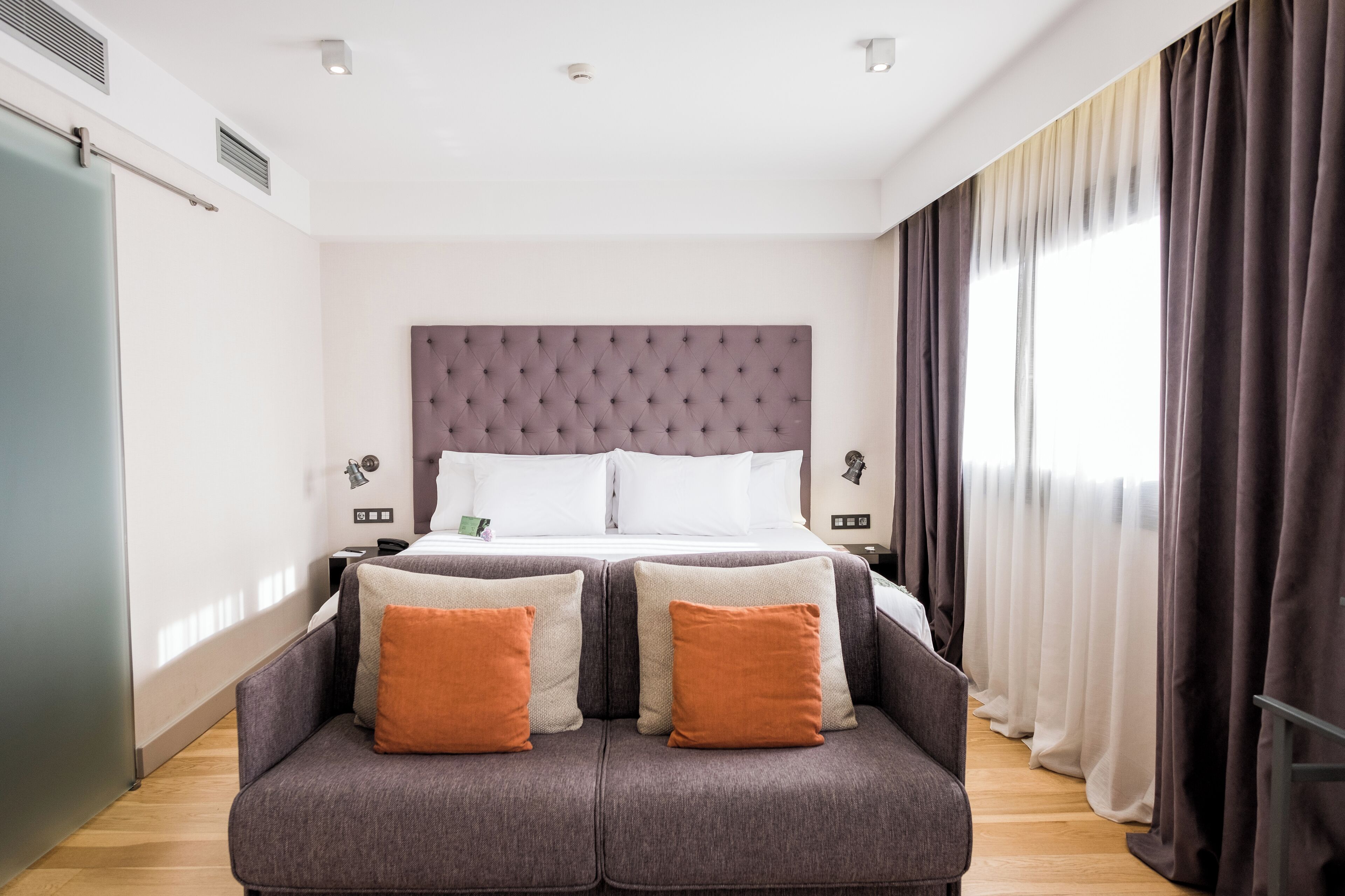 Una habitación de hotel moderna con una cama grande, dos mesitas de noche, un sofá con cojines y vistas al exterior a través de amplios ventanales.