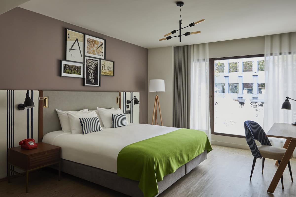 A hotel room with a large bed, a desk with a chair, a red telephone, and a window with curtains.